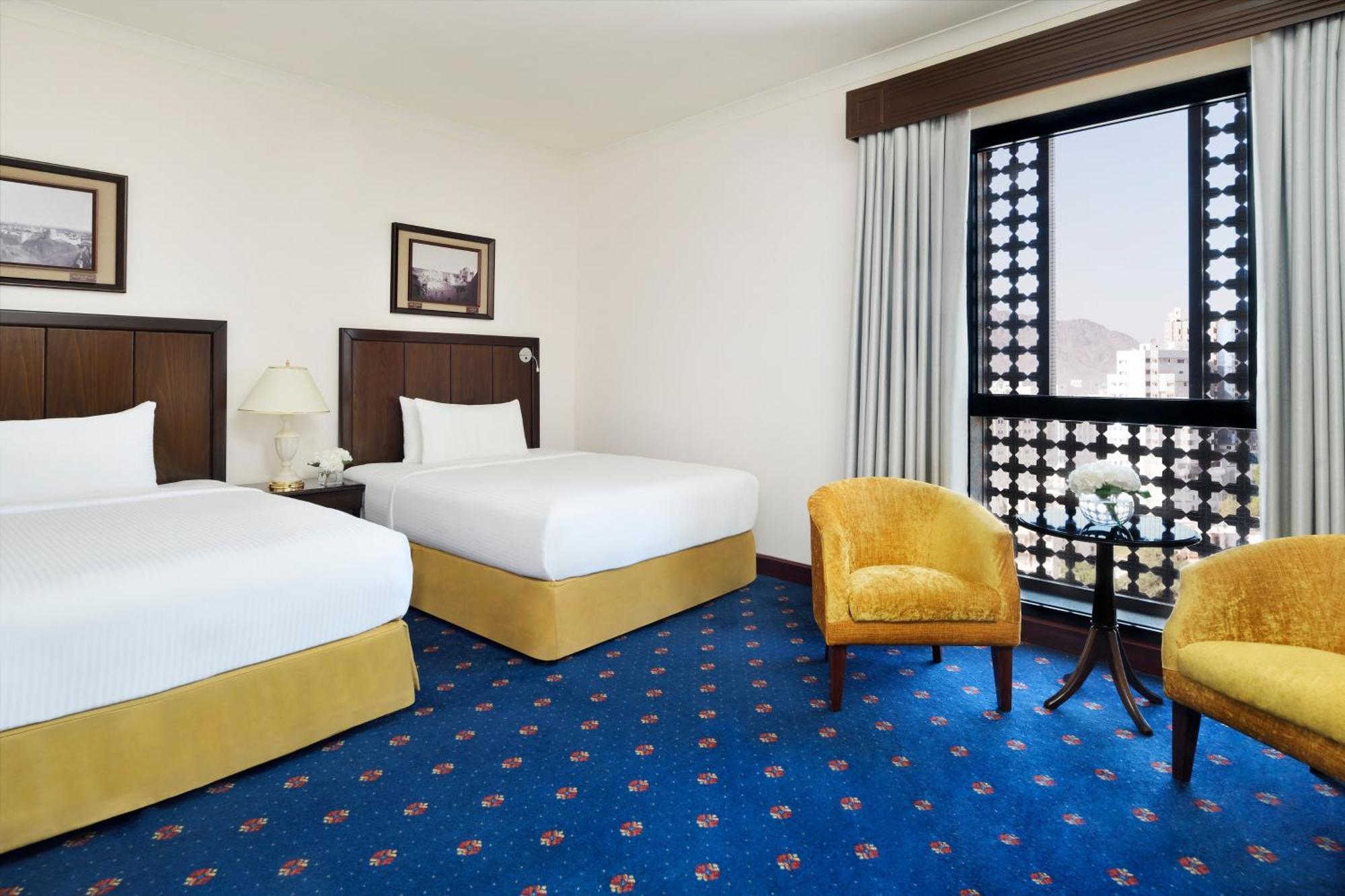 Intercontinental Dar Al Hijra Madinah, An Ihg Hotel Medina Exterior photo The photo shows a hotel room featuring two twin beds with crisp white linens and yellow bases. There are framed pictures on the walls above the beds. A small table and two yellow armchairs are positioned beside the window, which has decorative curtai