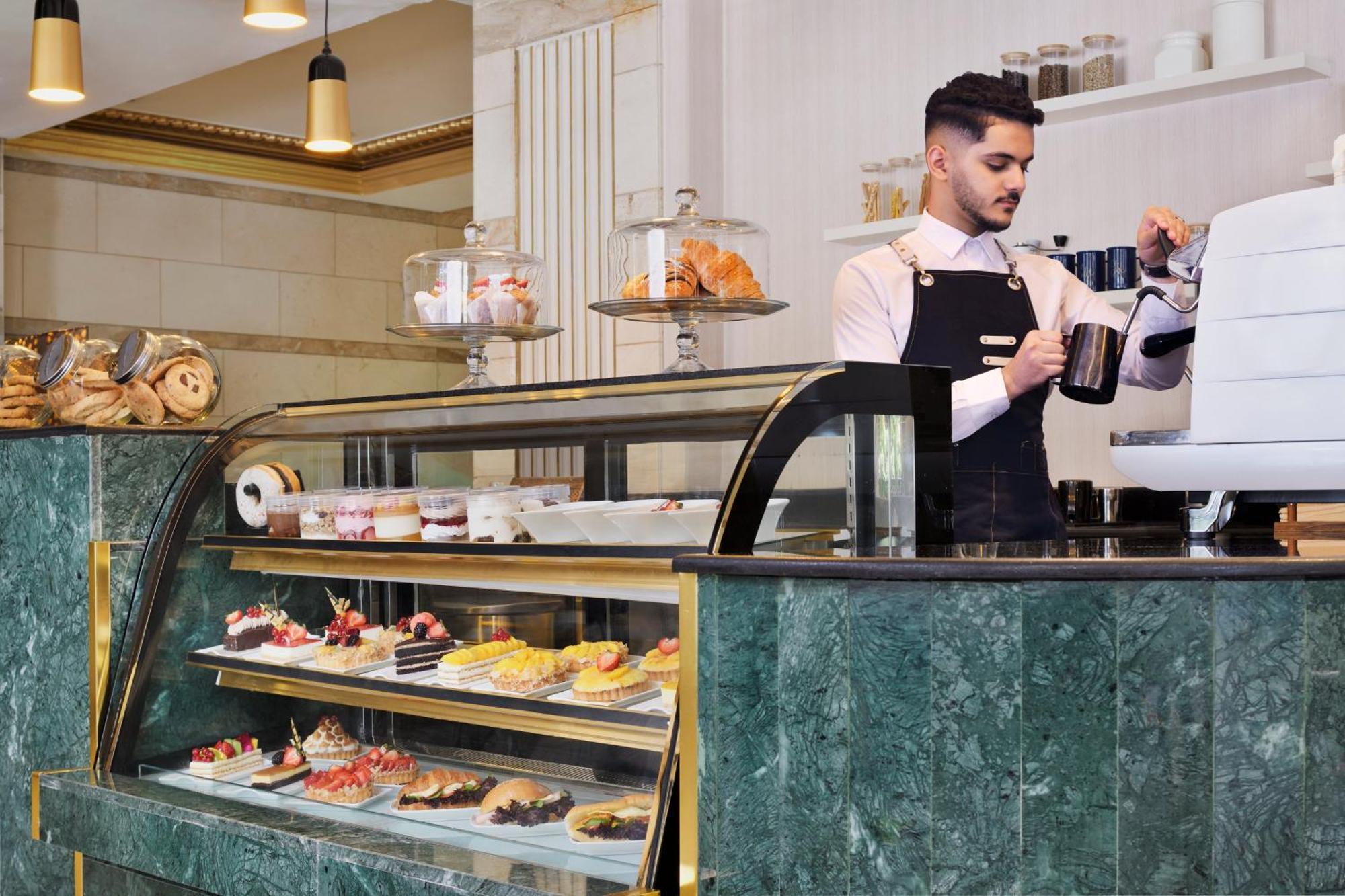 Intercontinental Dar Al Hijra Madinah, An Ihg Hotel Medina Exterior photo The photo shows a stylish bakery or café interior. In the foreground, there is a glass display case filled with a variety of pastries and desserts, showcasing colorful treats. Behind the counter, a barista wearing a white shirt and black apron is pre